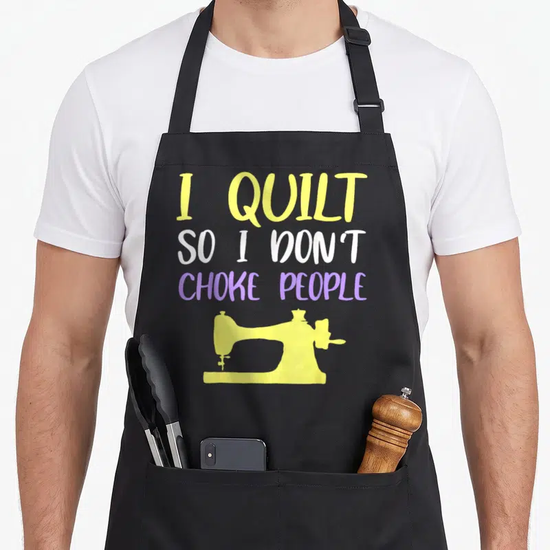 Choke quilt apron - I QUILT SO I DON'T CHOKE PEOPLE text in yellow white purple above sewing machine silhouette — shown on a black cotton canvas apron against a white background