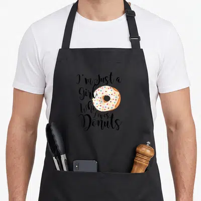 Vintage donut apron - Stacked black script I'm Just a Girl Who Loves Donuts around pink frosted donut with blue yellow — shown on a black cotton canvas apron against a white background