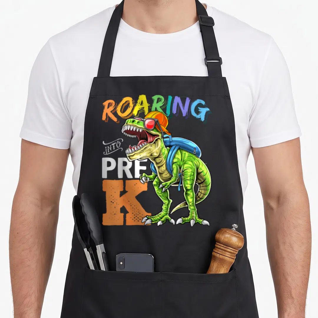Backpack dinosaur apron - Green T-Rex with orange cap blue backpack and ROARING INTO PRE K text — shown on a black cotton canvas apron against a white background
