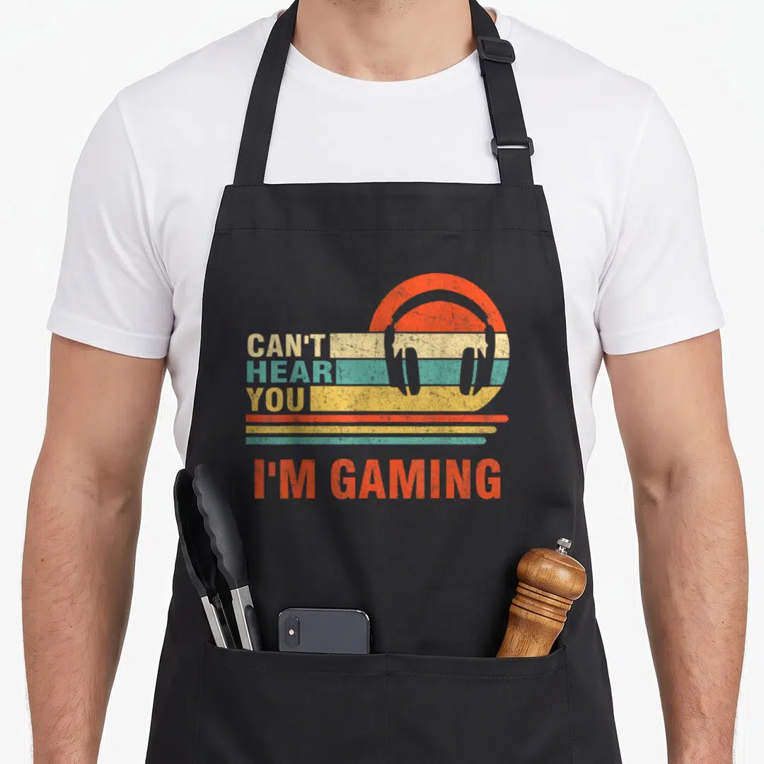 Gamer headset gaming apron - Retro striped circle with headphone graphic and stacked text CAN'T HEAR YOU I'M GAMING — shown on a black cotton canvas apron against a white background