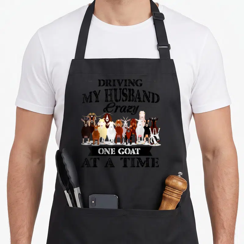 Husband goat driving aprons - Cartoon row of black white brown and tan goats with humorous text — shown on a black cotton canvas apron against a white background