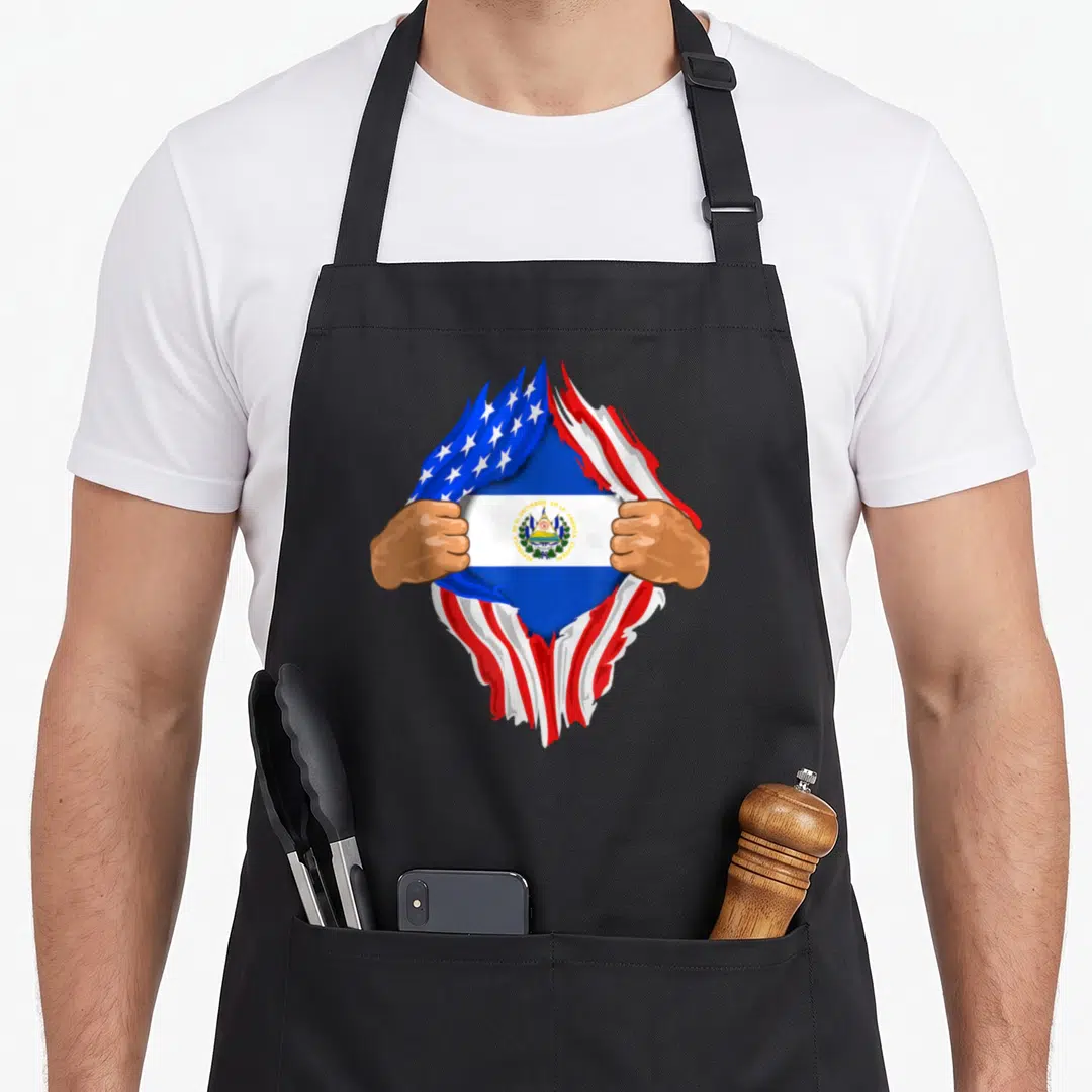 Blood salvadoran apron - Two brown hands tear a US flag to reveal El Salvador flag with Spanish text — shown on a black cotton canvas apron against a white background