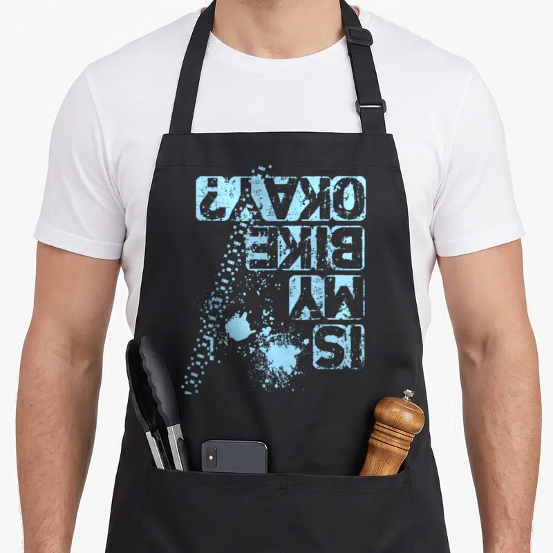 Retro bike apron - Light blue distressed text Is My Bike Okay rotated vertically with paint splatters — shown on a black cotton canvas apron against a white background