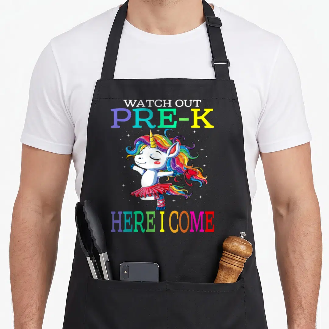 Watch unicorn apron - Cartoon unicorn in tutu with rainbow mane and WATCH OUT PRE-K HERE I COME text — shown on a black cotton canvas apron against a white background