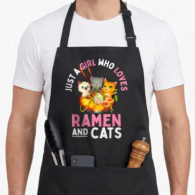 Ramen neko kawaii cat apron - Three cartoon cats in ramen bowl with pink text Just a Girl Who Loves Ramen and Cats — shown on a black cotton canvas apron against a white background