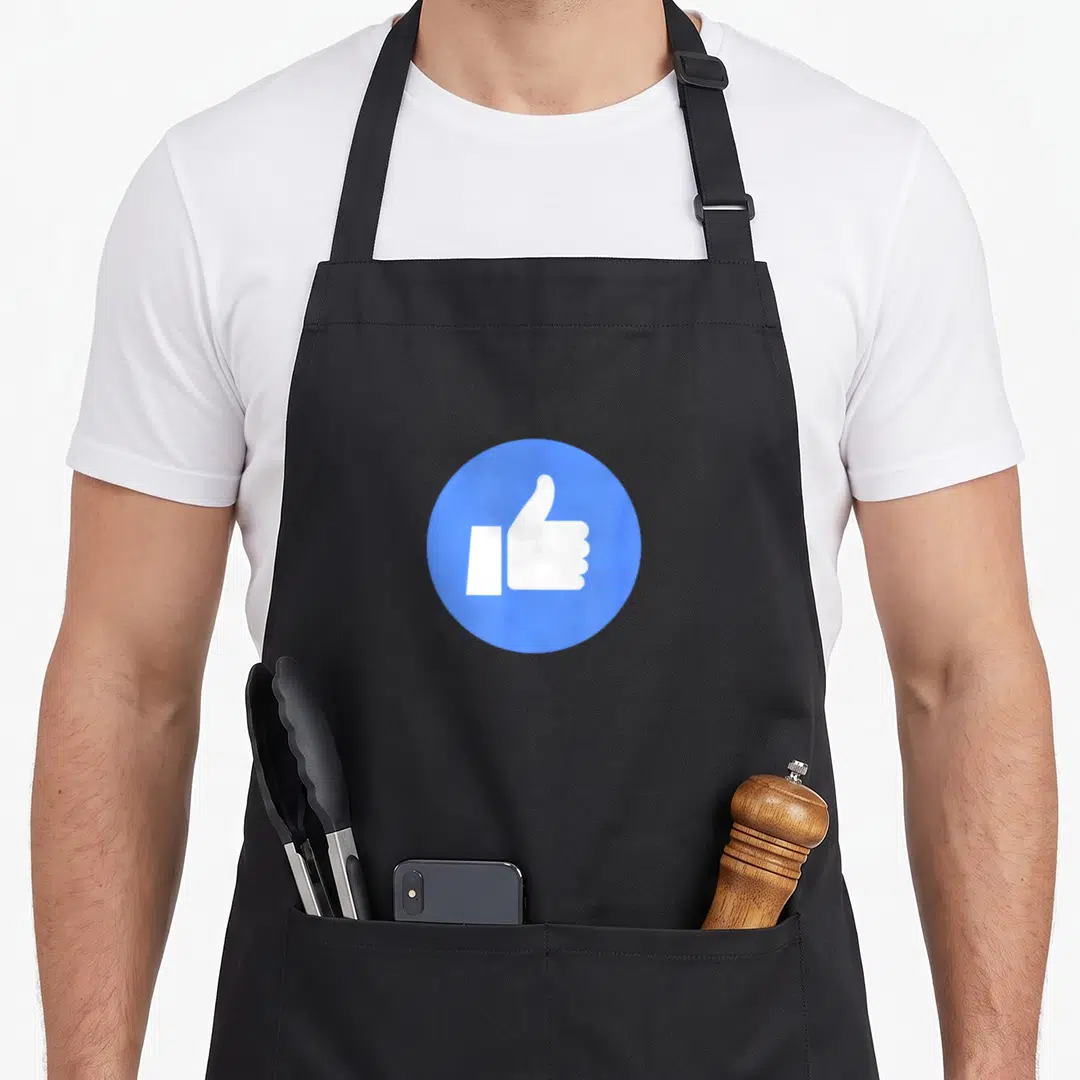 Thumb face apron - White thumbs up icon centered inside a solid blue circle on fabric — shown on a black cotton canvas apron against a white background