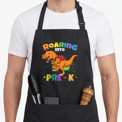 Roaring kindergarten boys apron - Orange T-Rex with red backpack, multicolor ROARING INTO PRE-K text, paint splatters — shown on a black cotton canvas apron against a white background