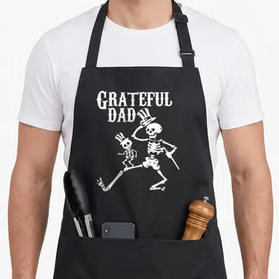 Grateful halloween apron - White distressed skeleton dad with top hat and cane holding child skeleton — shown on a black cotton canvas apron against a white background