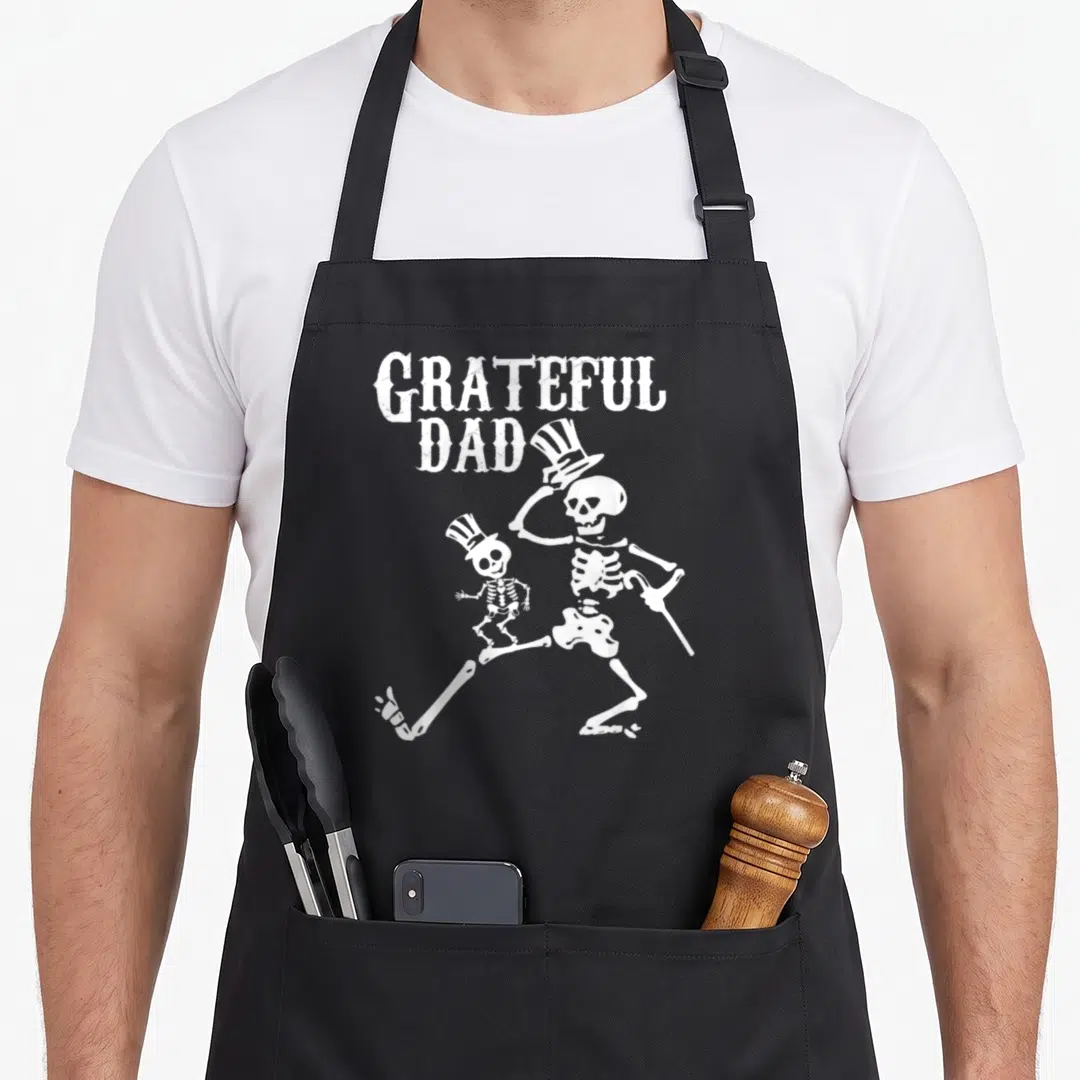 Grateful halloween apron - White distressed skeleton dad with top hat and cane holding child skeleton — shown on a black cotton canvas apron against a white background