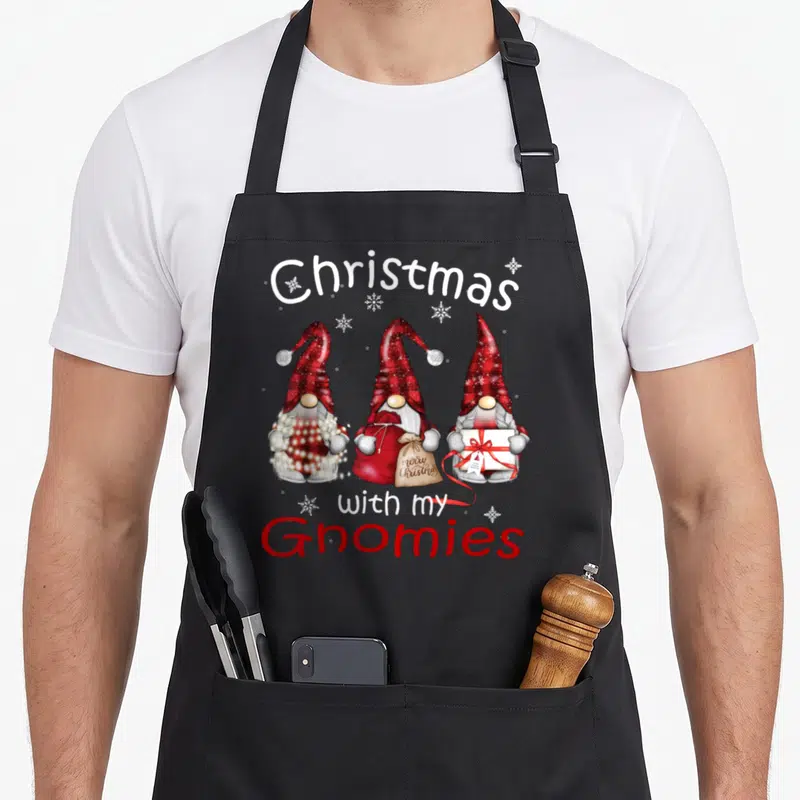 Gnome buffalo apron - Three red-hatted gnomes holding gifts under Christmas with my Gnomies text — shown on a black cotton canvas apron against a white background