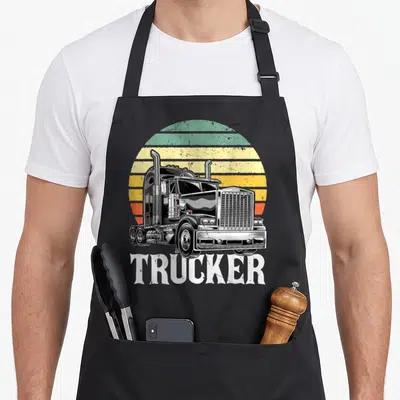 Trailer driver semi truck apron - Retro semi-truck illustration with striped sun and TRUCKER text in black white yellow — shown on a black cotton canvas apron against a white background