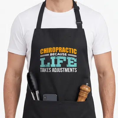 Chiropractor student aprons - Orange and blue text reading CHIROPRACTIC BECAUSE LIFE TAKES ADJUSTMENTS with white lines — shown on a black cotton canvas apron against a white background