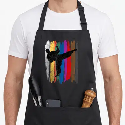 Belt taekwondo apron - Black martial artist kicking silhouette over vertical white, yellow, blue, purple, red, brown — shown on a black cotton canvas apron against a white background