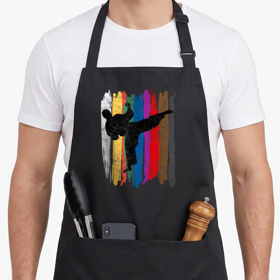 Belt taekwondo apron - Black martial artist kicking silhouette over vertical white, yellow, blue, purple, red, brown — shown on a black cotton canvas apron against a white background