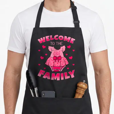 Gender baby shower apron - Pink cartoon baby dress with bow and hearts surrounded by welcome to the family text — shown on a black cotton canvas apron against a white background