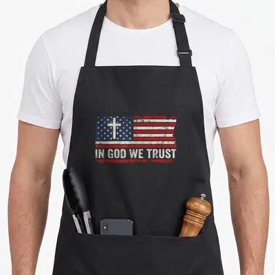 God patriotic apron - Distressed USA flag with white cross in blue field and IN GOD WE TRUST text on red stripe — shown on a black cotton canvas apron against a white background