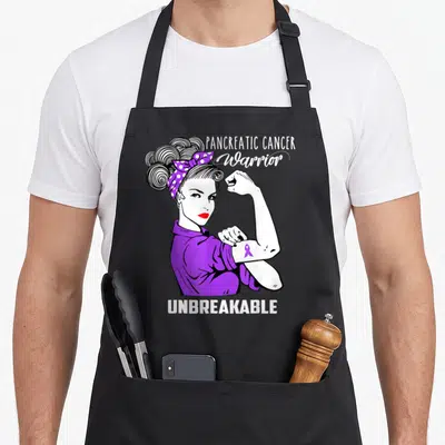 Pancreatic cancer awareness apron - Cartoon woman flexing purple ribbon with PANCREATIC CANCER Warrior UNBREAKABLE text — shown on a black cotton canvas apron against a white background