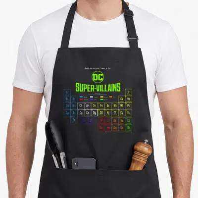 Villain table apron - DC Super-Villains periodic table grid with colored location boxes and character symbols — shown on a black cotton canvas apron against a white background