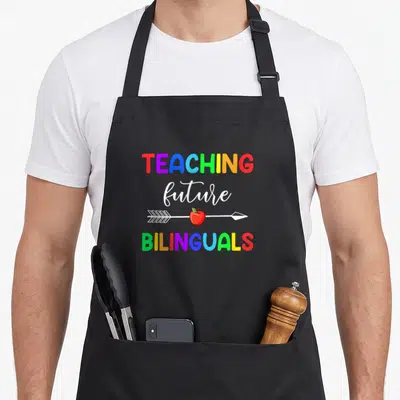 Teaching spanish aprons - Rainbow text TEACHING future BILINGUALS with arrow and red apple on black background — shown on a black cotton canvas apron against a white background