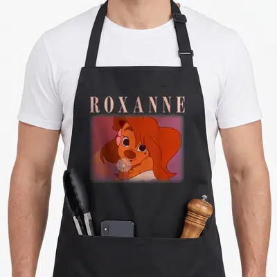 Movie valentine apron - Roxanne cartoon portrait with name text above head on blurred pink background — shown on a black cotton canvas apron against a white background