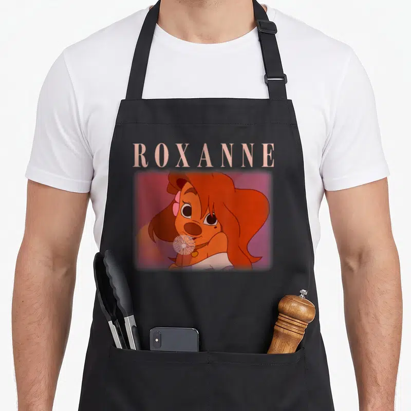 Movie valentine apron - Roxanne cartoon portrait with name text above head on blurred pink background — shown on a black cotton canvas apron against a white background