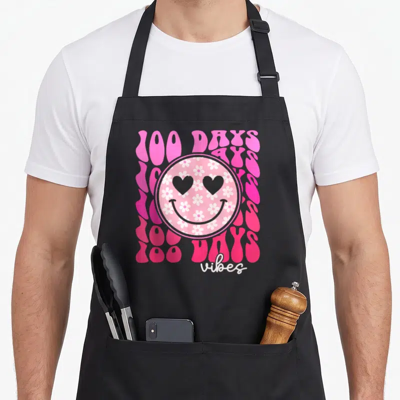 100th daisy apron - Retro pink smiley face with white flower pattern and stacked 100 DAYS text — shown on a black cotton canvas apron against a white background