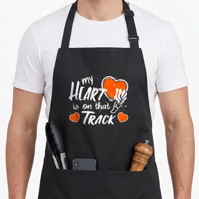 Field track apron - White text my HEART is on that TRACK with orange heart and running shoe — shown on a black cotton canvas apron against a white background