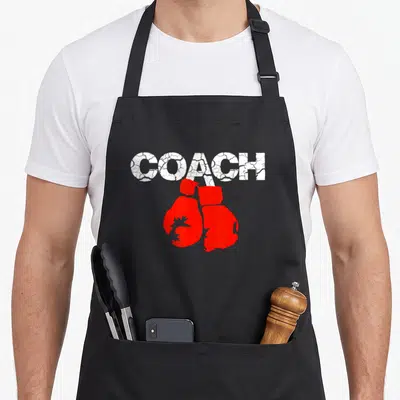 Definition boxing coach apron - Distressed white COACH text above a pair of red boxing gloves — shown on a black cotton canvas apron against a white background