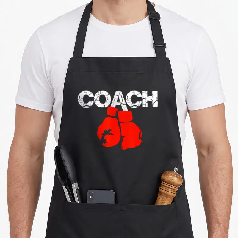 Definition boxing coach apron - Distressed white COACH text above a pair of red boxing gloves — shown on a black cotton canvas apron against a white background