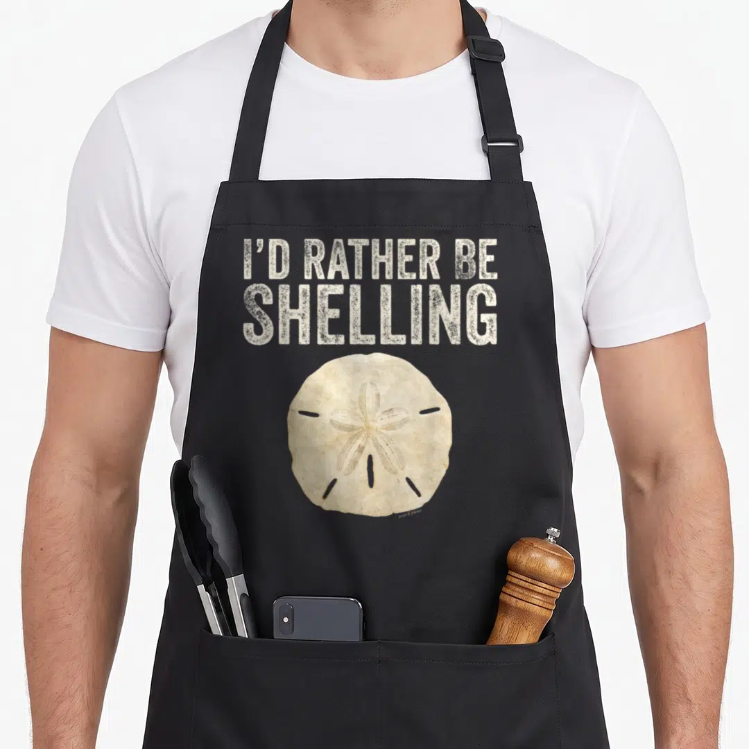 Rather hunter apron - Distressed I'D RATHER BE SHELLING text above a beige sand dollar illustration — shown on a black cotton canvas apron against a white background