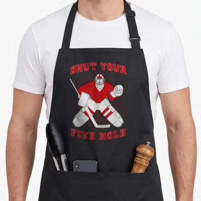 Shut hockey dad apron - Cartoon goalie in red gear with text SHUT YOUR FIVE HOLE arched above and below — shown on a black cotton canvas apron against a white background