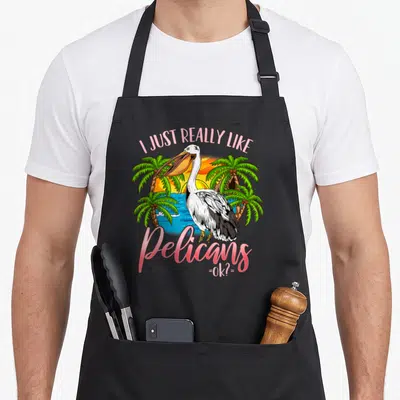 Funny pelican apron - Cartoon pelican on tropical beach with pink text I Just Really Like Pelicans =Ok?= — shown on a black cotton canvas apron against a white background
