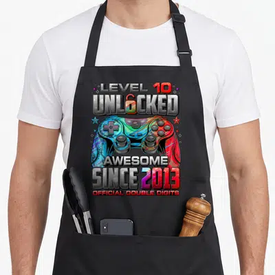 10th gaming aprons - LEVEL 10 UNLOCKED text above a blue and red game controller with stars and 2013 date — shown on a black cotton canvas apron against a white background