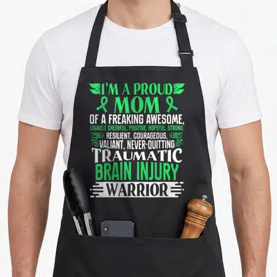 Traumatic ribbon apron - Stacked green and white text reading proud mom traumatic brain injury warrior with ribbons — shown on a black cotton canvas apron against a white background