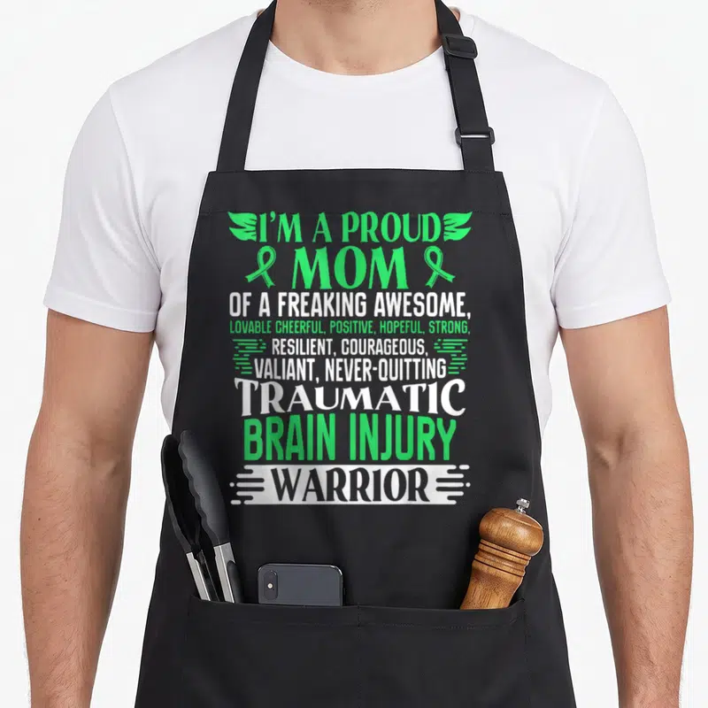 Traumatic ribbon apron - Stacked green and white text reading proud mom traumatic brain injury warrior with ribbons — shown on a black cotton canvas apron against a white background