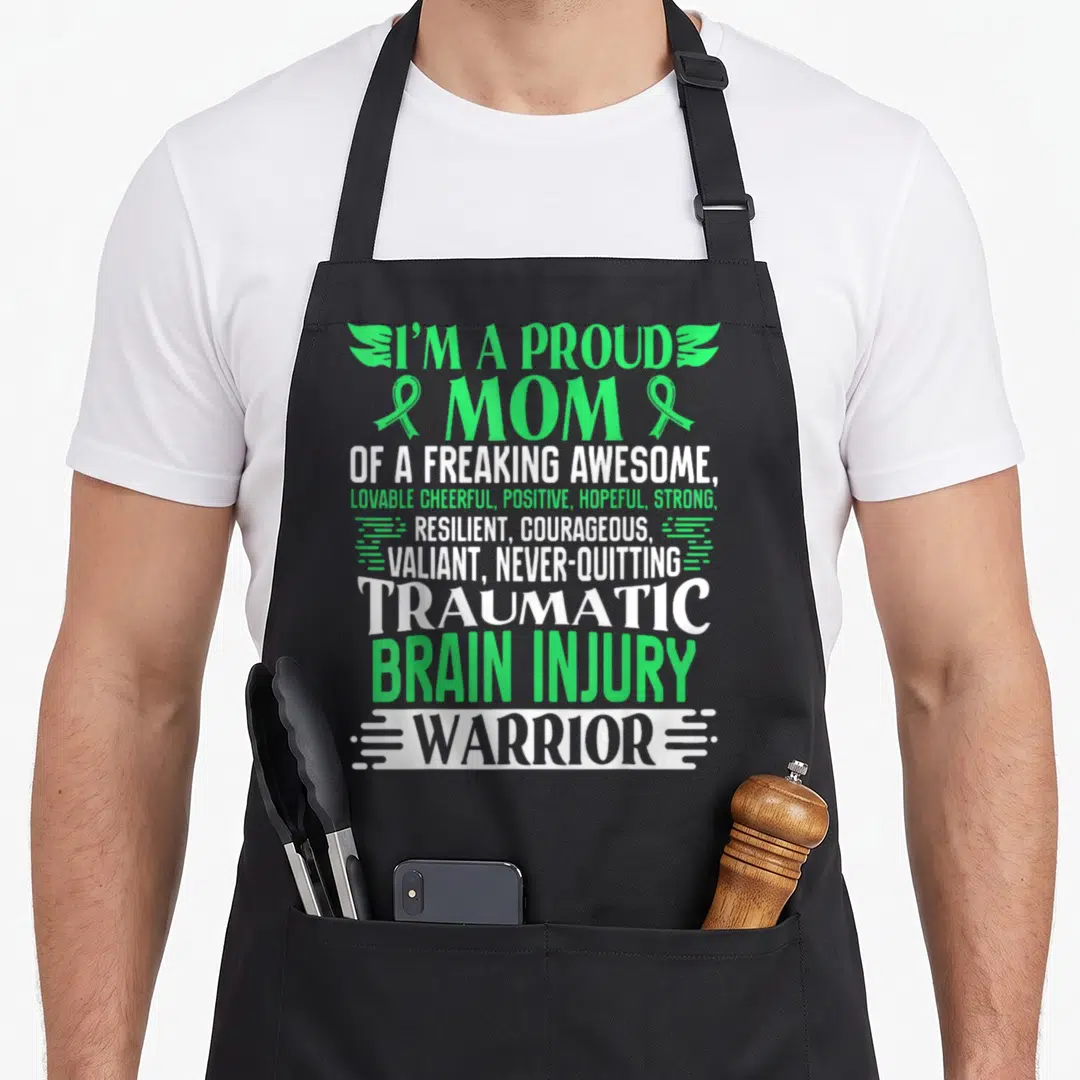 Traumatic ribbon apron - Stacked green and white text reading proud mom traumatic brain injury warrior with ribbons — shown on a black cotton canvas apron against a white background