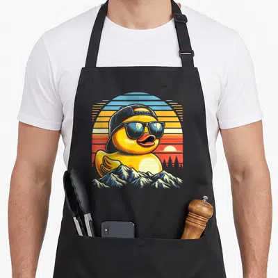 Duck rubber apron - Yellow cartoon duck with cap and sunglasses in front of retro sunset and mountains — shown on a black cotton canvas apron against a white background