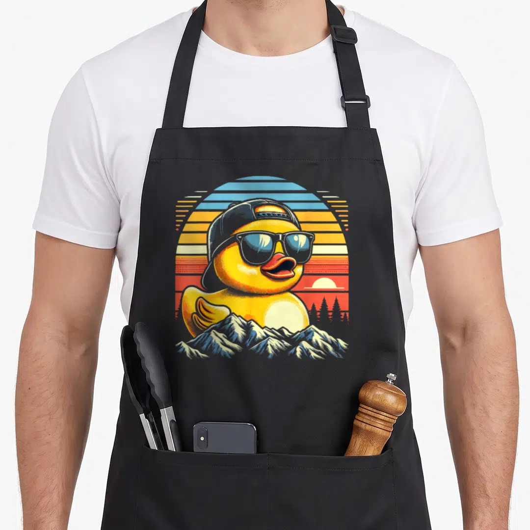 Duck rubber apron - Yellow cartoon duck with cap and sunglasses in front of retro sunset and mountains — shown on a black cotton canvas apron against a white background