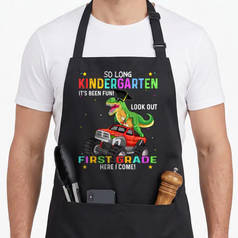 Class dinosaur apron - Green T-Rex in red monster truck with graduation cap and colorful school text — shown on a black cotton canvas apron against a white background