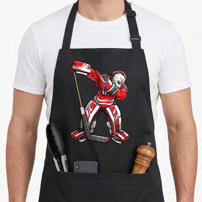 Dabbing hockey apron - Cartoon hockey goalie in red and white gear holding a stick in a dynamic pose — shown on a black cotton canvas apron against a white background
