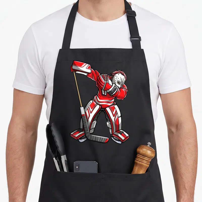 Dabbing hockey apron - Cartoon hockey goalie in red and white gear holding a stick in a dynamic pose — shown on a black cotton canvas apron against a white background