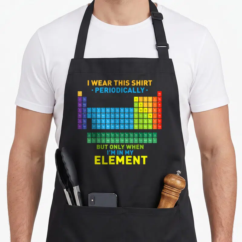 Periodic table chemistry apron - Colorful periodic table chart with yellow blue green red and purple blocks and pun text — shown on a black cotton canvas apron against a white background