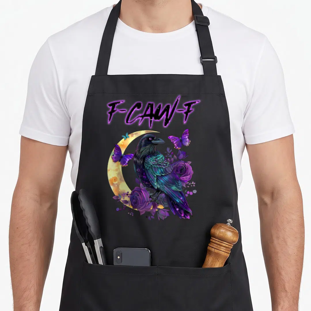 Caw raven halloween apron - F-CAW-F text above a crow perched on purple roses and blue butterflies against a yellow — shown on a black cotton canvas apron against a white background