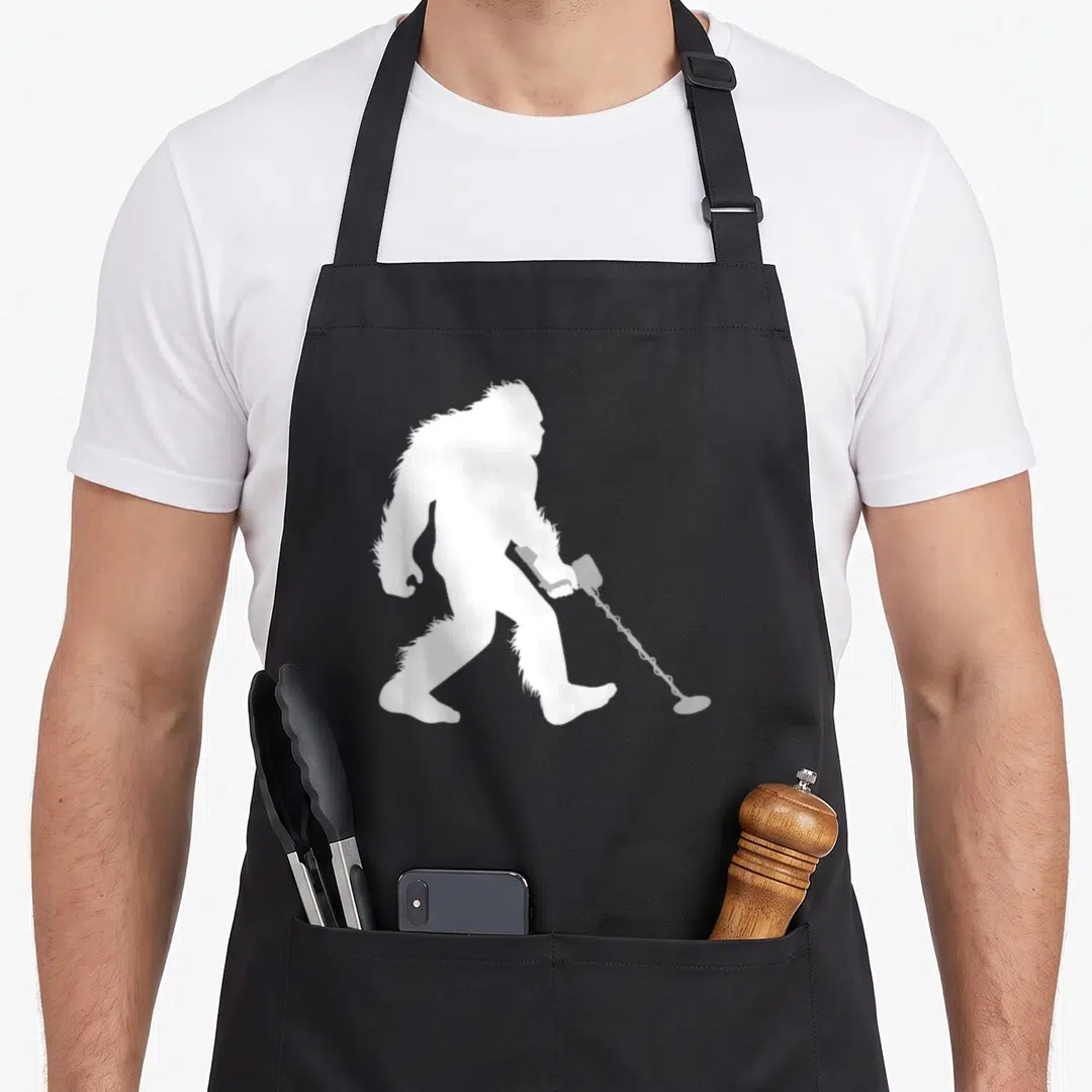 Bigfoot metal detecting apron - White silhouette of bigfoot walking right holding a metal detector — shown on a black cotton canvas apron against a white background