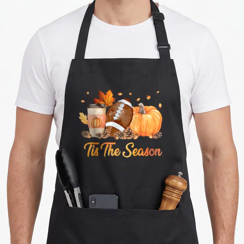 Tis fall apron - Watercolor football, pumpkin, coffee cup, pinecones, and oak leaves above orange script reading Tis — shown on a black cotton canvas apron against a white background