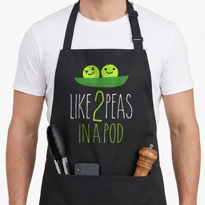 Peas vegetable apron - Cartoon two peas in a pod with green and white text reading LIKE 2 PEAS IN A POD — shown on a black cotton canvas apron against a white background