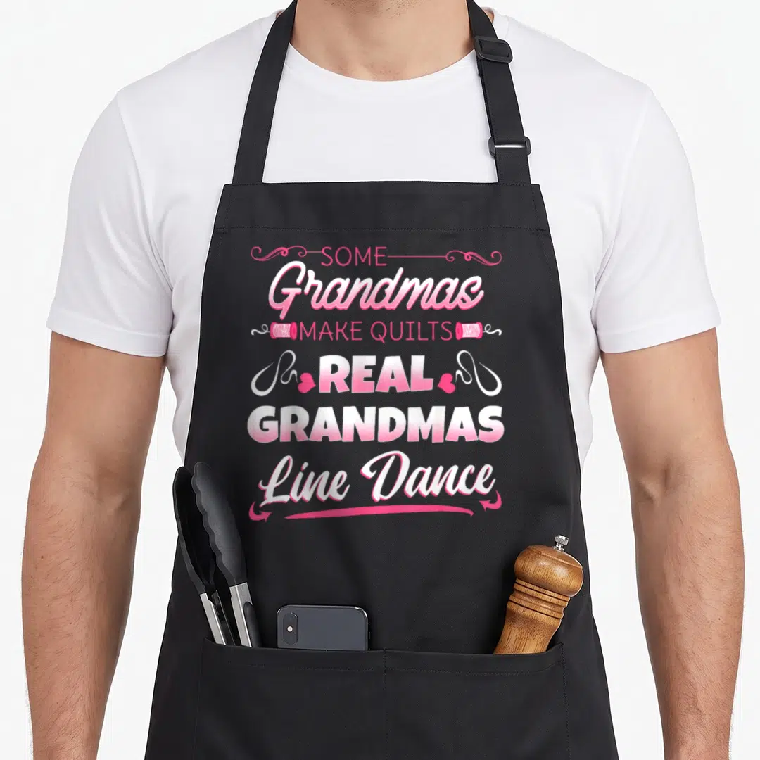 Quilt grandma's aprons - Stacked pink and white text reading Some Grandmas Make Quilts Real Grandmas Line Dance — shown on a black cotton canvas apron against a white background