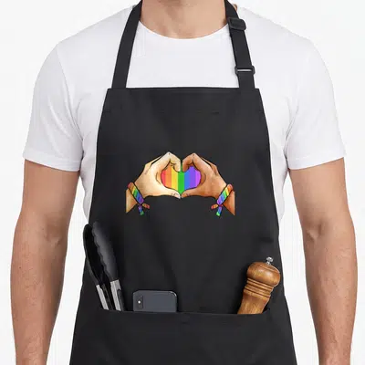 Lgbt gay pride apron - Two tan cartoon hands form a heart filled with rainbow stripes and matching wristbands — shown on a black cotton canvas apron against a white background