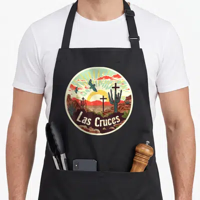 Cross mexico apron - Circular Las Cruces desert scene with sun, mountains, crosses, and birds in red, yellow, teal — shown on a black cotton canvas apron against a white background