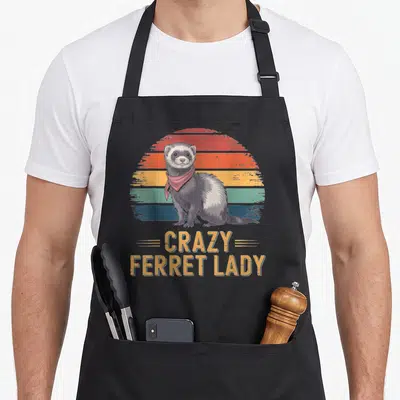 Vintage ferret animal apron - Retro ferret in red bandana before striped sun with stacked CRAZY FERRET LADY text — shown on a black cotton canvas apron against a white background